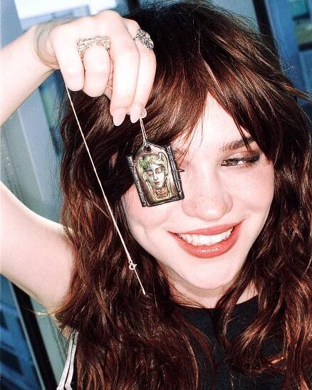 a woman holding up a sculpture of a face that is on a chain, she's covering her left eye with it and is smiling