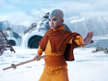 aang in the live action netflix avatar the last airbender. a young boy wearing red and yellow loose clothing, holding a wooden staff, and with a blue arrow tattooed on his forehead