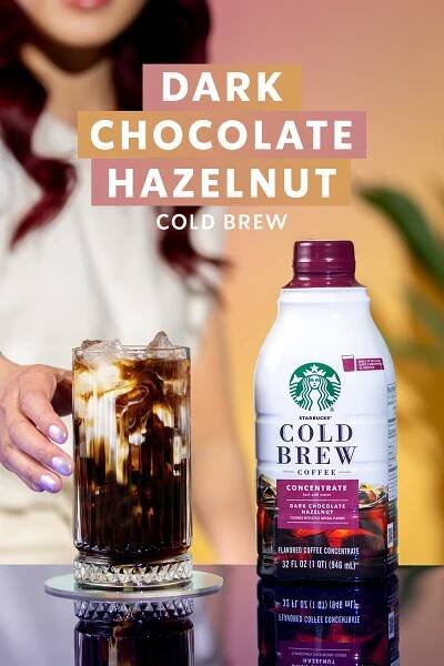 A woman reaches for a glass of cold brew coffee with creamer on a table. Next to it is a bottle of Starbucks Dark Chocolate Hazelnut Cold Brew Concentrate.