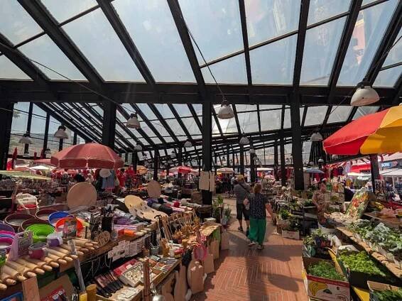 A bustling indoor market with glass roof, stalls selling goods like baskets, vegetables, wooden items, and household products. Shoppers walk between displays under red and yellow umbrellas. Bright, busy, and vibrant scene.