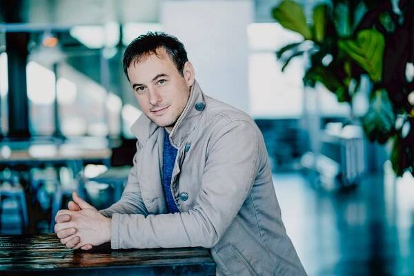 A white man wearing a light jacket leaning on a wooden table