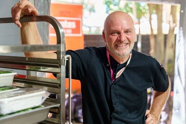 Cheerful person in black polo shirt smiling while standing next to shelving or equipment in what appears to be a shop.