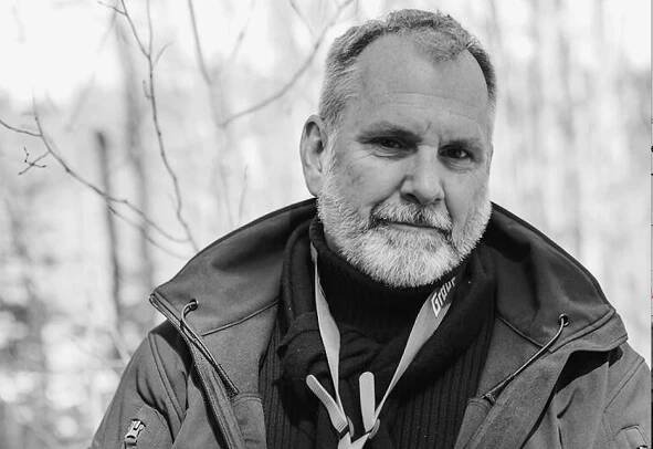 an older bearded man in a heavy coat stands in front of spindly trees.