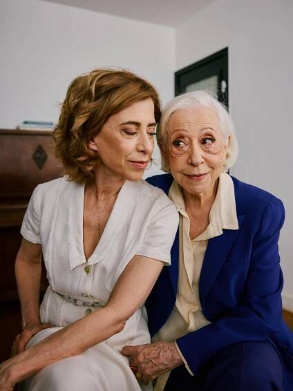 A woman in a white dress sits close to an older woman wearing a blue blazer.