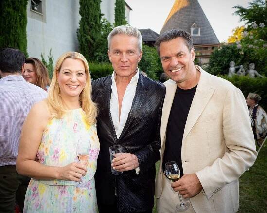 Daniela Winkler, Alfons Haider und Didi Tunkel stehen mit Weingläsern lachend im Schlossgarten.