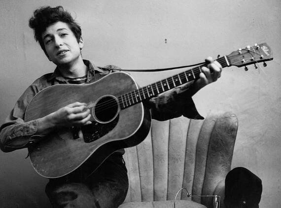 A young bob dylan smiling and playing a gibson acoustic guitar while sitting on the arm of a chair
