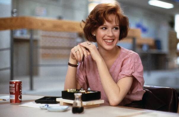 Molly Ringwald in The Breakfast Club with short hair smiles at a table with a meal and a drink