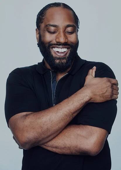 John David Washington wears a navy top and hugs himself, smiling wide.
