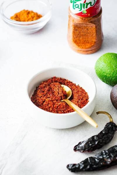 A homemade spice blend of chilli flakes, citric acid, dehydrated limes and salt in a small white bowl with a bottle of Tajín Clásico Seasoning in the background.