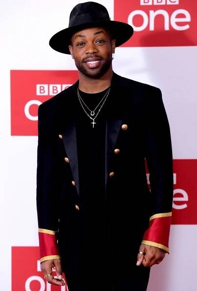 Todrick Hall channels bold elegance in a black military-inspired blazer with gold buttons and red cuff accents, layered over a black tee and accessorized with layered necklaces and a wide-brimmed hat at a BBC One event