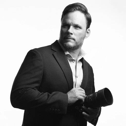 A man wearing a suit holds a camera, looking off to the side. Black and white photograph.