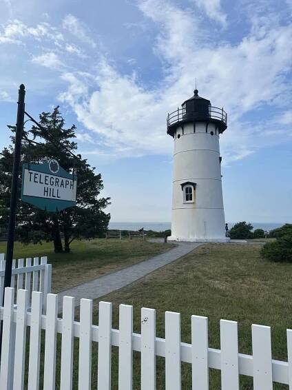 A white lighthouse with a black top stands on a grassy hill under a blue sky with scattered clouds. A sign reads "Telegraph Hill" near a white picket fence, and the ocean is visible in the background.