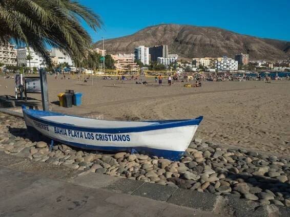 Los Cristianos, Tenerife, Kanárské ostrovy, Španělsko, 2. ledna 2022: Pohled na písečnou pláž Playa de Los Cristianos s modrobílou dřevěnou lodí, opalování turistů, holé kopce a apartmány. — Stock obrázek
