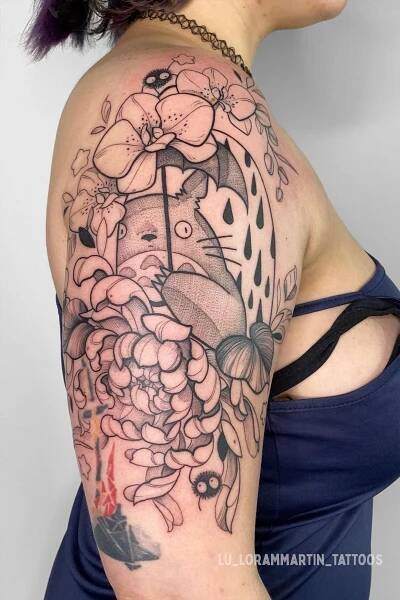 Shows an arm with fresh black linework tattoo of a peony and Totoro under an umbrella, surrounded by soot sprites, inspired by Studio Ghibli's My Neighbor Totoro. With black line details and shading.