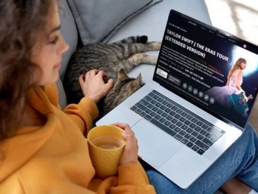 A woman sitting next to a cat with a laptop on her lap watching Taylor Swift The Eras Tour Concert.