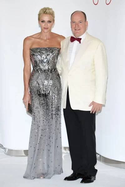 A couple posing elegantly; woman in silver sequin gown and man in white tuxedo with a bow tie on a white backdrop.