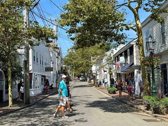A lively street scene with pedestrians walking along a tree-lined road surrounded by white clapboard buildings. Storefronts with American flags and hanging flower baskets create a picturesque small-town atmosphere.