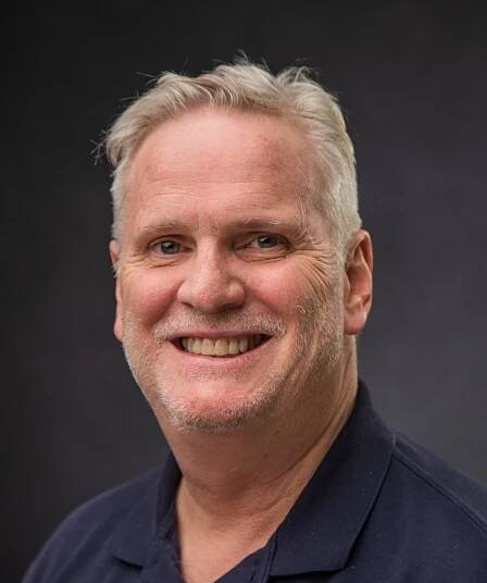 Smiling middle-aged man with short light gray hair and light stubble, wearing a navy blue collared shirt, posing against a dark, plain background.