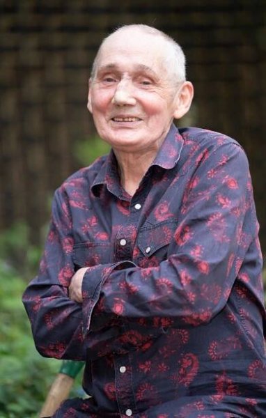 an elderly man wearing a red and black shirt smiles with his arms crossed
