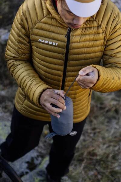 Person wearing a yellow Mammut jacket and cap compressing a gray outdoor gear bag.