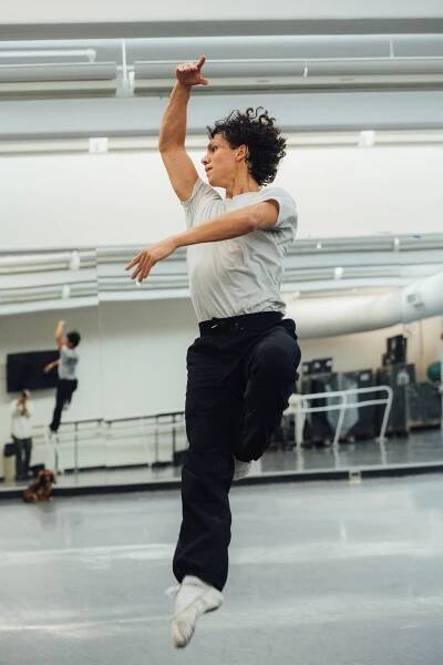 A ballet dancer in white T-shirt and black pants, his curly hair flopping, jumps in a studio, one arm curved around his head, his body reflected in a mirror behind him.