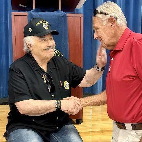 A warm interaction between two men, one wearing a black shirt with accessories including a cross necklace and sunglasses on his head, and the other in a red polo shirt. They are shaking hands, conveying a friendly connection in a setting with a blue backdrop.