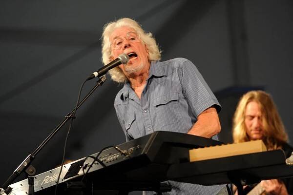 John Mayall, a man with long white hair and a beard, singing into a microphone and playing a keyboard onstage.