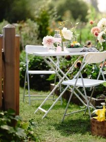 V rozkvetlé zahradě jsou dvě židle a stůl TORPARÖ s růžovými nápoji, vázami a mísou salátu.