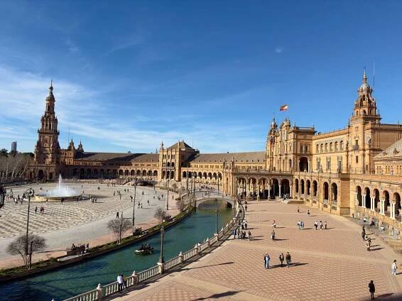 The image features the Plaza de España in Seville, Spain, a must-see among things to do in Sevilla. It showcases its semi-circular building, central fountain, and a canal with small boats. People are walking and enjoying the sunny day as the Spanish flag flies atop one of the towers.