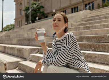 Stáhnout - A beautiful young girl is enjoying a delicious coffee while sitting on the stairs. Attractive female model in a striped blouse is satisfied with a drink. — Stock obrázek