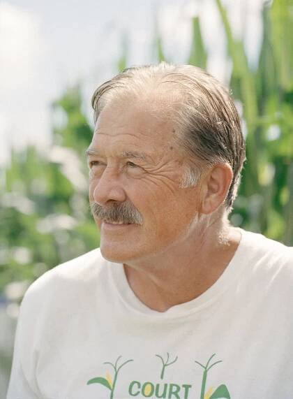 A close-up portrait shows Mark Kuhn, with a mustache and partially gray hair, standing in front of out-of-focus cornstalks.