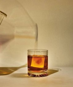 photo of brown liquor poured from beaker into highball glass with large square ice cube