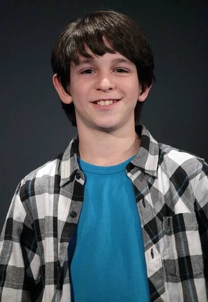 A person smiling, wearing a plaid shirt over a blue T-shirt, standing against a plain backdrop