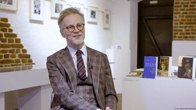 Jacek Dehnel, seated and smiling, in front of a table with books. He has glasses and a neat white beard; he is wearing a smart three-piece suit in a brown check, with a brown patterned tie.