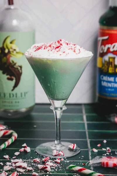 A festive peppermint Christmas mocktail sits elegantly in a martini glass, crowned with whipped cream and crushed peppermint. Bottles of spirits and candy pieces create a cheerful display on the green countertop.