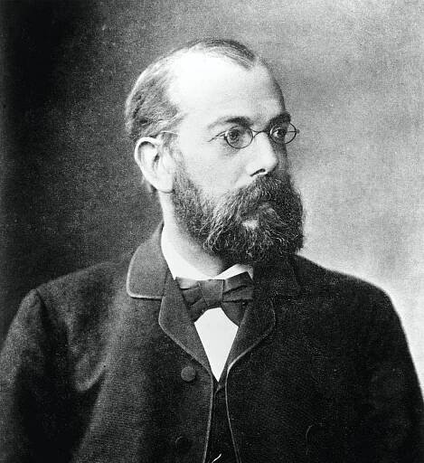 Black and white portrait of a middle-aged bearded man with a light skin tone wearing small wireframe glasses and a suit with a bowtie