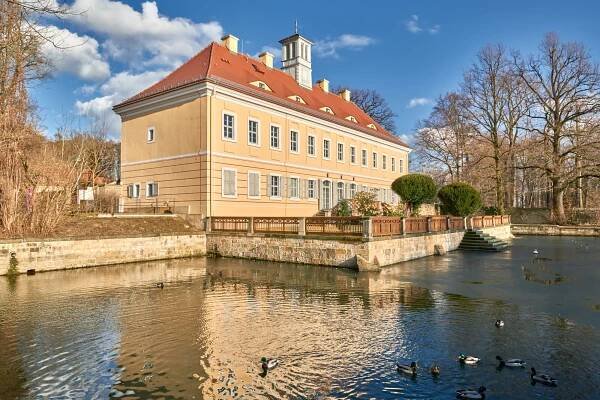 Ein großes beiges Gebäude mit rotem Dach steht neben einem ruhigen Teich mit Enten. Das Gebäude ist von einer Steinterrasse umgeben und die Bäume sind jetzt kahl, was auf Winter hindeutet. Der Himmel ist klar und blau mit ein paar vereinzelten Wolken.
