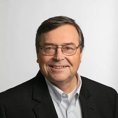 A man with glasses and graying hair wearing a dark suit jacket and a light striped shirt smiles at the camera.