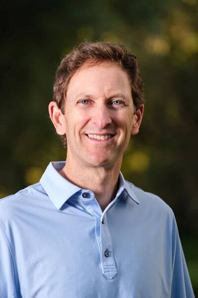 Corporate headshot of a man dressed in a blue collared shirt, standing against a natural outdoor background.