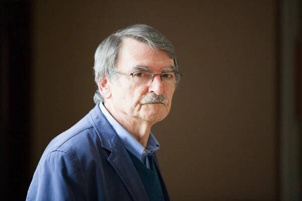 A close-up portrait of him indoors standing  in sunlight. He has gray hair and a mustache, wears half-rimmed eyeglasses and is turning his head to look at the camera. He has on a blue jacket over a blue shirt. 