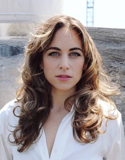 Photo a woman with long, layered brown hair captured during a portrait photo shoot. She appears in an outdoor setting, dressed in fashionable clothing. The focus is on her face and neck, highlighting her features as she poses for the camera.