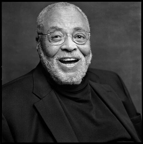 An elderly person with a beard smiles warmly, wearing glasses and a dark suit with a turtleneck
