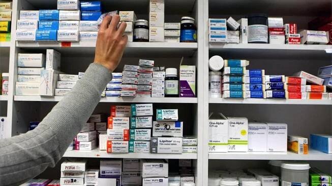 A woman's arm is stretching up to a shelf of a unit stacked with medicines in boxes and bottles.