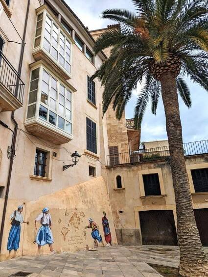 A vibrant street scene in Palma, Mallorca, featuring historical buildings with large windows and a mural depicting figures in traditional attire next to a tall palm tree. The mural adds a touch of local culture to the quiet, sunlit square.