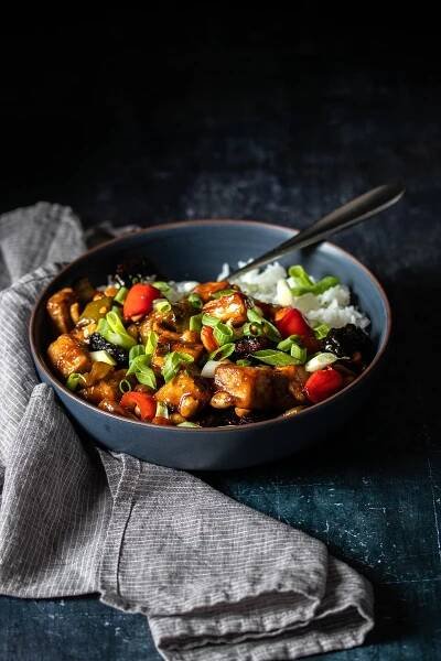 A dark blue bowl on a dark moody background with kung pao tofu and peppers topped with green onions.