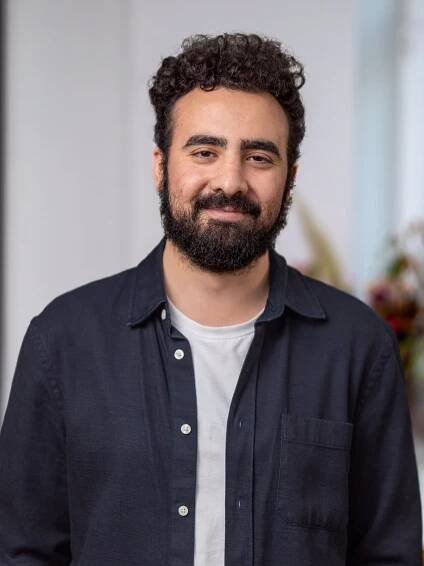 Ein Mann mit lockigem dunklem Haar, einem gepflegten Bart und einem warmen Lächeln steht in einem sanft beleuchteten Innenraum. Er trägt ein marineblaues Hemd mit Knopfleiste über einem weißen T-Shirt, was für einen entspannten und dennoch professionellen Look sorgt. Sein freundlicher Gesichtsausdruck und sein leicht geneigter Kopf vermitteln Zugänglichkeit. Der Hintergrund ist verschwommen und bietet ein modernes Ambiente mit subtilen floralen Elementen, das an ein modernes Büro oder eine kreative Umgebung erinnert.