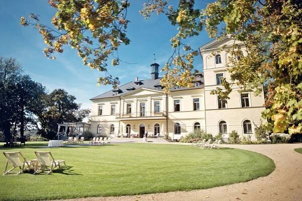A grand mansion, Chateau Mcely, surrounded by lush green lawn and tall trees, exuding elegance and charm.