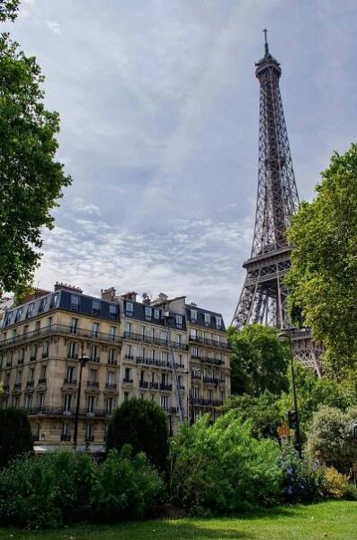 The Eiffel Tower rises behind classic Parisian apartment buildings, partly framed by tall trees and lush greenery. The sky is partly cloudy, casting soft light over the scene. The photo captures a quiet neighborhood feel near one of the city’s most iconic landmarks.