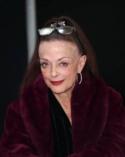 A dark-haired woman in a burgundy coat and a pair of glasses sitting on top of her head.
