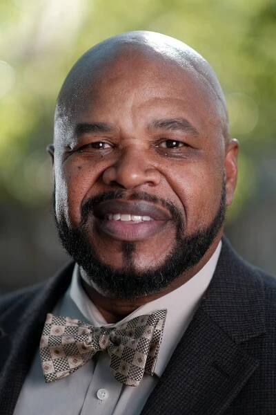 Professional headshot of a man with a beard and a shaved head, wearing a tailored suit and a bowtie, photographed in an outdoor environment.